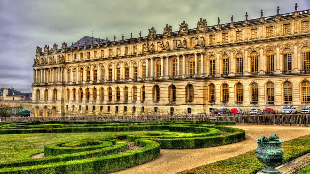 Exterior view of the Palace of Versailles with manicured gardens in the foreground and a cloudy sky above.
